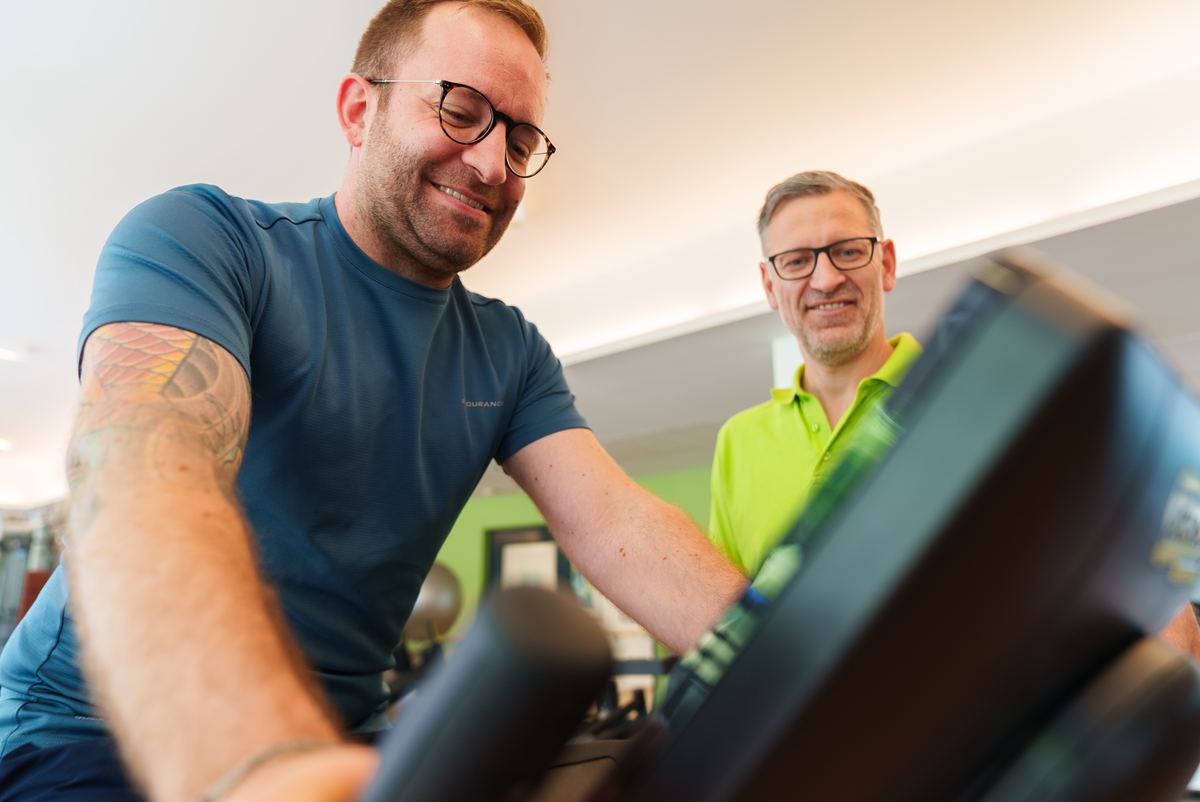 Therapeut im grünen Poloshirt betreut einen Patienten am Trainingsgerät, beide lachen